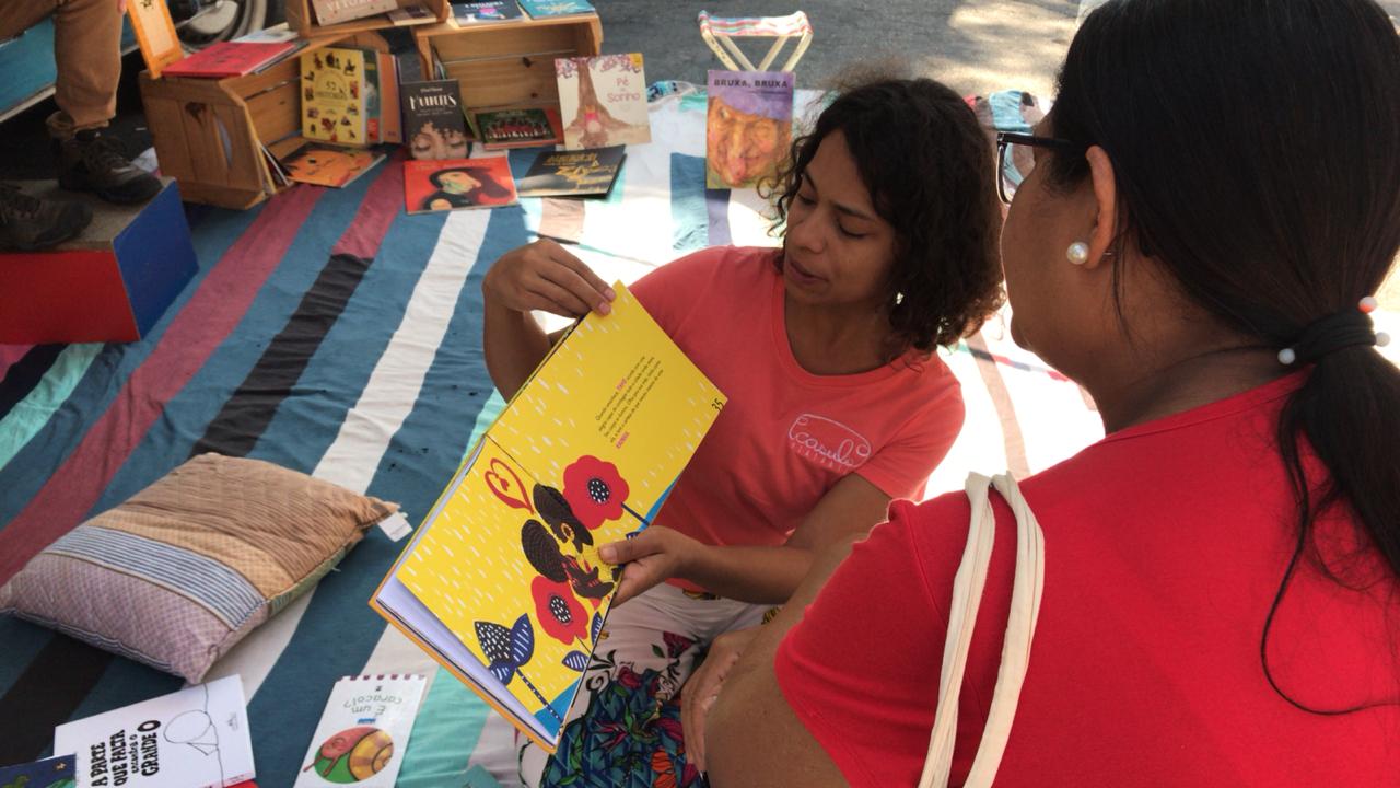 Foto em ambiente bastante colorido e alegre, com almofadas e tecido listrado ao chão e destaque para duas mulheres sendo uma delas segurando um livro e mostrando para a outra